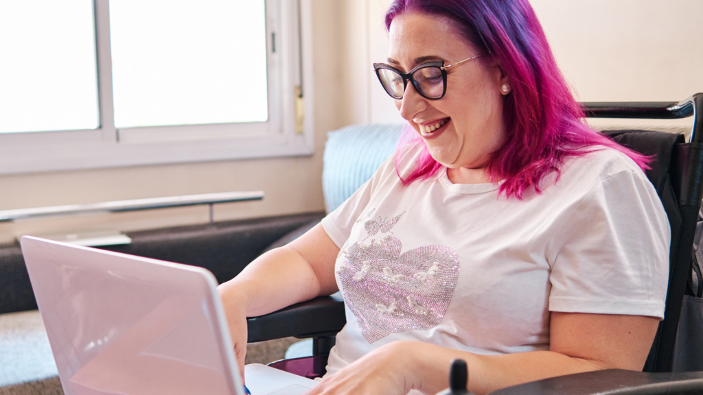 Woman in wheelchair with pink hair smiling and using laptop